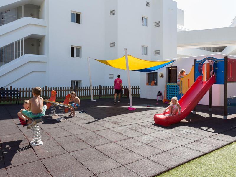 Děti si hrají na venkovním hřišti s červenou skluzavkou a houpačkou.