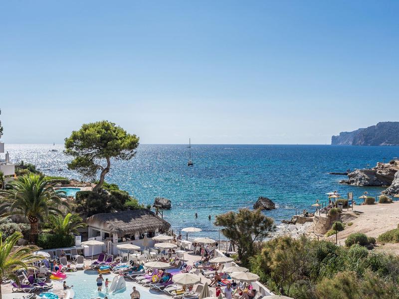 Živá pláž s bazénem, slunečníky a výhledem na moře a útesy.