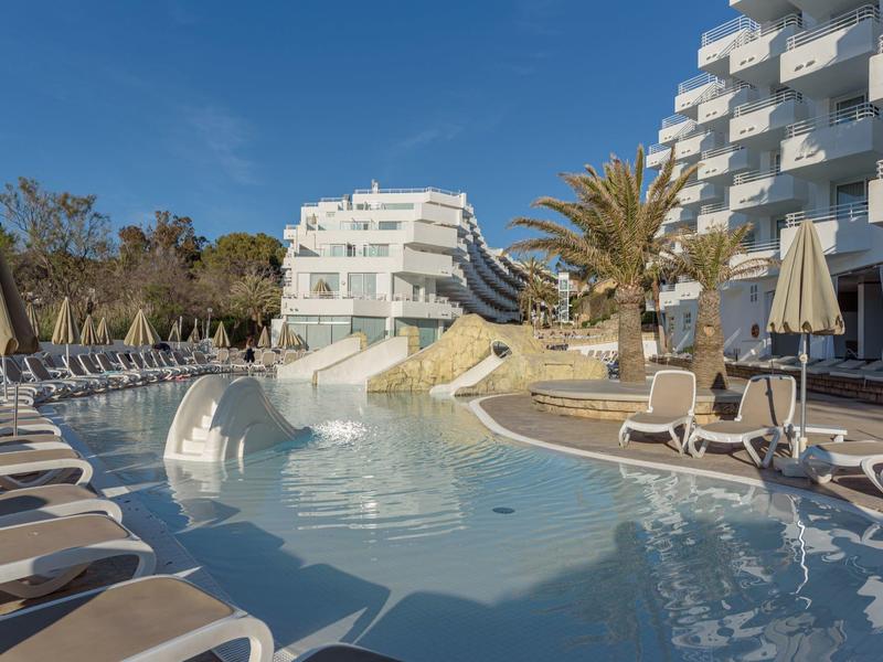 Hotelový bazén s lehátky a slunečníky pod modrou oblohou.