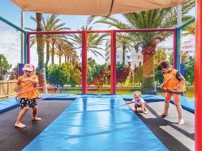 Kinder springen und spielen auf einem großen rechteckigen Trampolin im Freien bei Sonnenschein.