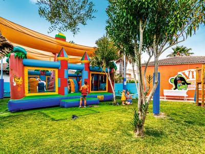 Bunte Hüpfburg mit Kindern auf grüner Wiese, umgeben von Bäumen und einem orangen Gebäude.