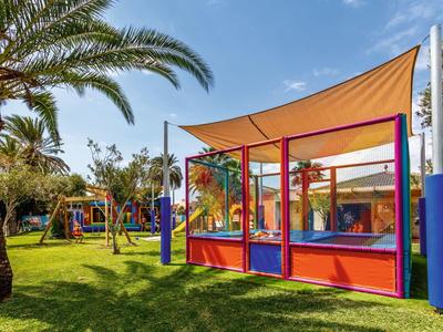 Kinderspielplatz mit buntem Klettergerüst, Schaukel, Rasenfläche und Palme unter blauem Himmel.