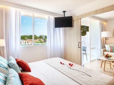 Helles, modernes Schlafzimmer mit großem Fenster, Doppelbett, roten Kissen und Wandfernseher.