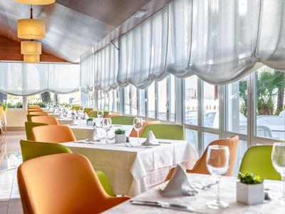 Modernes Restaurant mit großen Fenstern, weißen Tischdecken und orange-grünen Stühlen.