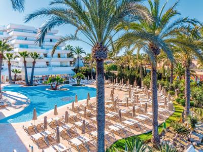 Großer blauer Pool mit Sonnenliegen und Palmen in hellem, sonnigem Resort-Außenbereich.