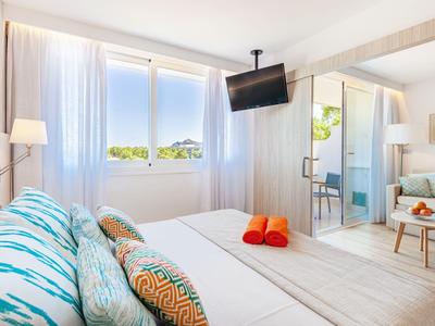 Helles Hotelzimmer mit großem Bett, bunten Kissen, Fernseher an der Decke und Balkon mit Meerblick.