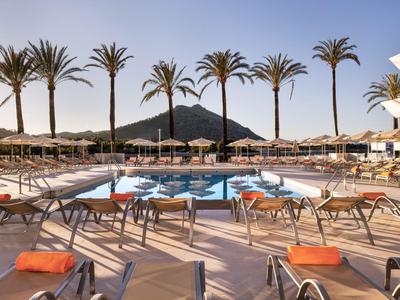 Espace piscine d'hôtel avec transats, palmiers et montagne en arrière-plan sous ciel clair.