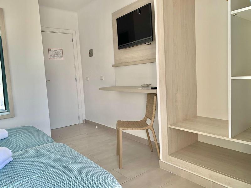 Minimalistische hotelkamer met twee bedden, wand-tv, houten stoel en open kastruimte.