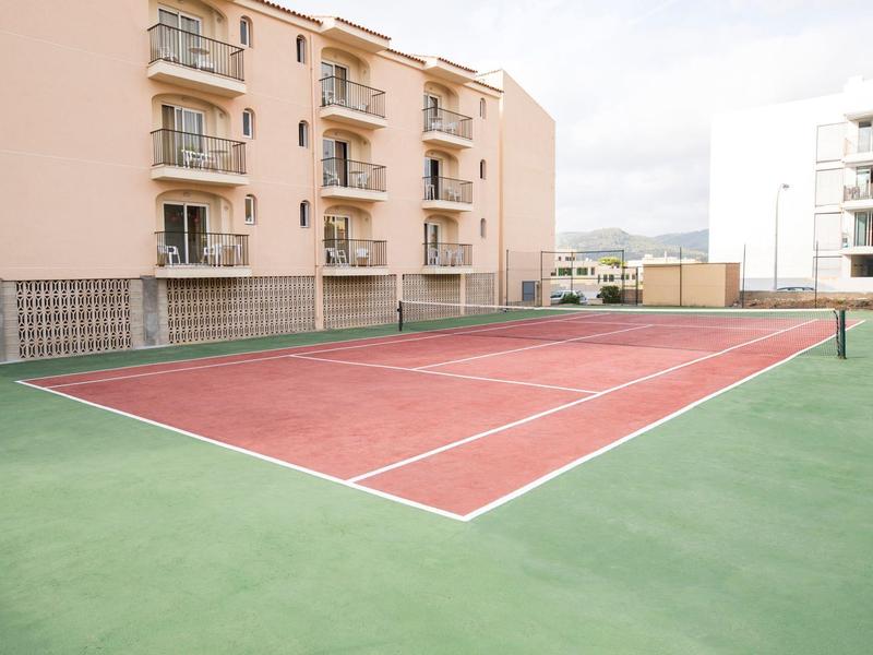 Buiten tennisbaan met rood-groene ondergrond naast appartementenbouw bij bewolkte lucht.