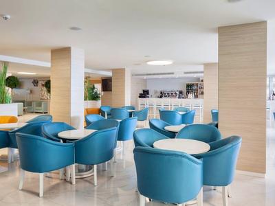Moderne Lounge mit blauen Sesseln, weißen Tischen, hellen Säulen und offener Bar im Hintergrund.