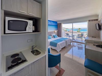 Kleines Hotelzimmer mit Kochnische, Doppelbett, Balkon und Meerblick, helle Farben und moderne Möbel.
