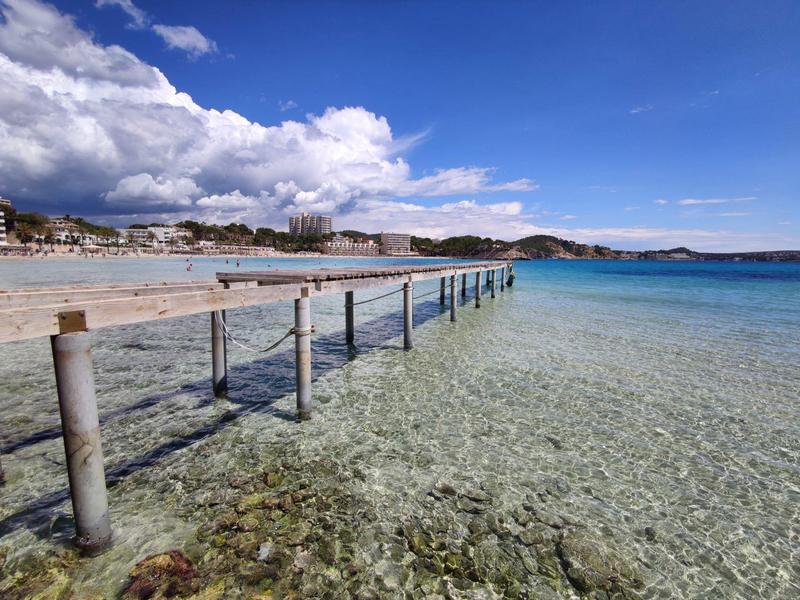 Long wooden pier over clear sea with sandy beach and cloudy sky in the background.