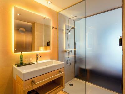 Modernes Badezimmer mit beleuchtetem Spiegel, Holzwaschtisch und Glas-Duschkabine mit Milchglasscheibe.