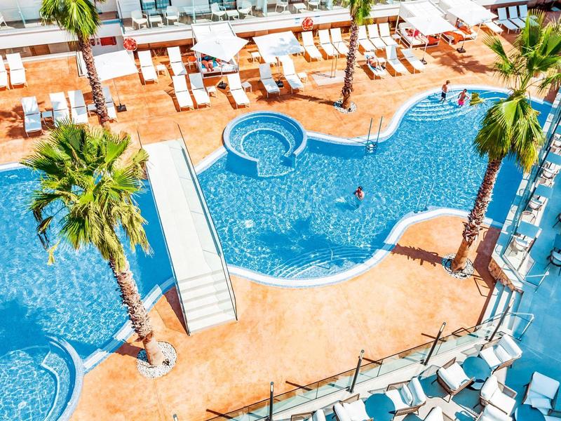 Piscina azul con tumbonas blancas y palmeras en un área de hotel soleada.