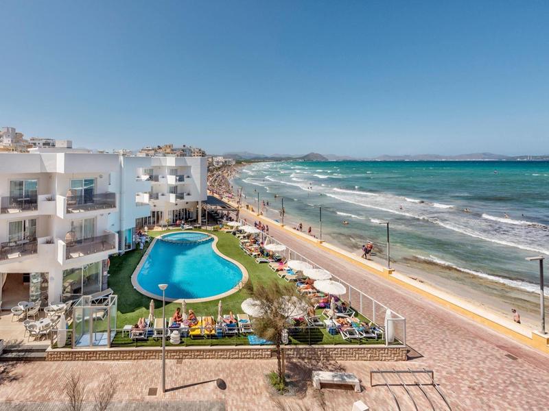 Kusthotel met een gebogen zwembad, promenade, zandstrand en golven onder een heldere lucht.