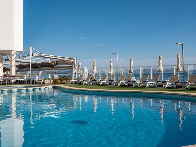 Buitenzwembad van hotel met ligstoelen onder parasols en heldere blauwe lucht.