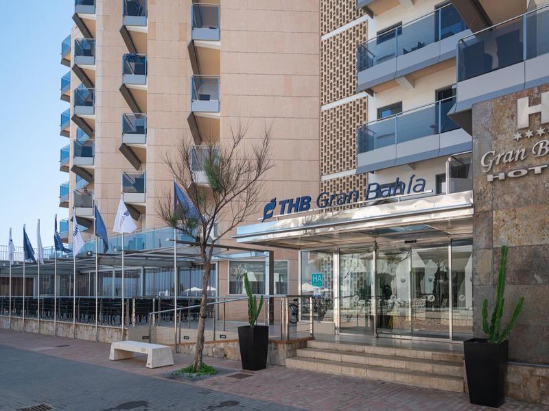 Moderne hotelentree met glazen deuren, balkons en een stenen gevel onder een heldere lucht.