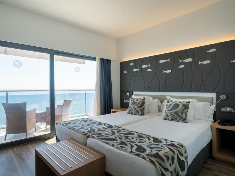 Moderne hotelkamer met tweepersoonsbed, decoratieve kussens, balkon en uitzicht op zee.