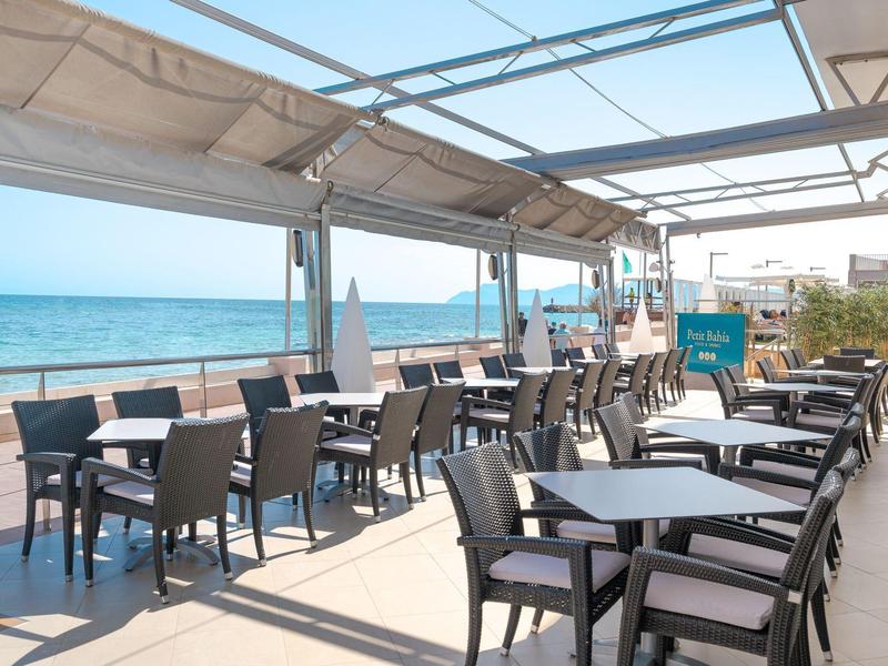 Buiten eetgedeelte met tafels, stoelen en zeezicht onder een overdekt terras bij een hotel.