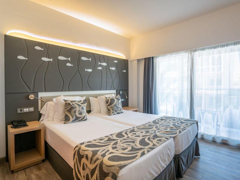 Lichte hotelkamer met aparte bedden, beddengoed met patroon, houten nachtkastje en groot raam met gordijnen.