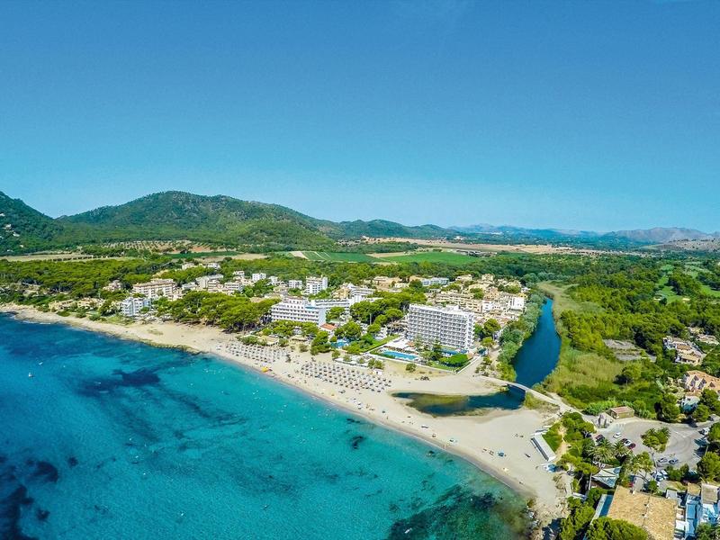 Littoral avec mer bleue, plage de sable et hôtels sous des collines boisées au ciel clair.