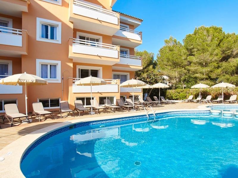 Piscine ronde avec chaises longues et parasols à côté d'un bâtiment hôtelier sous un ciel bleu.