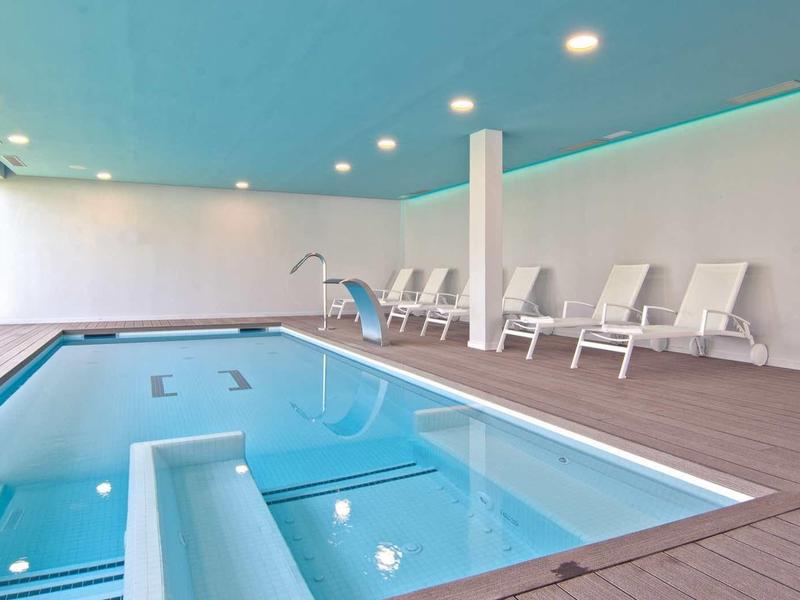 Nowoczesny kryty basen z białymi leżakami po jednej stronie, dużo naturalnego światła.