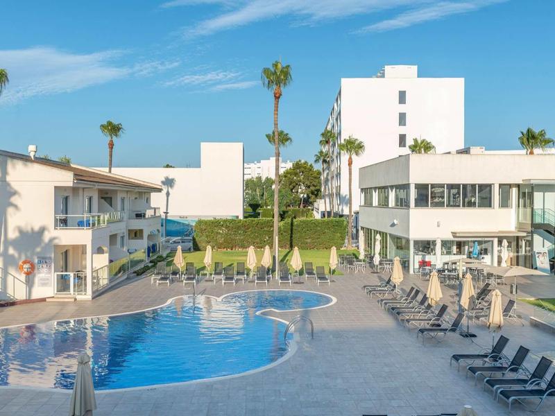 Zewnętrzny basen z leżakami między nowoczesnymi białymi budynkami pod niebieskim niebem.