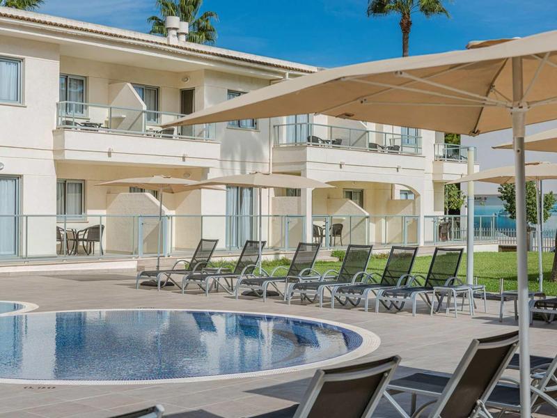 Hotel z okrągłym basenem, leżakami i dużymi parasolami pod jasnoniebieskim niebem.