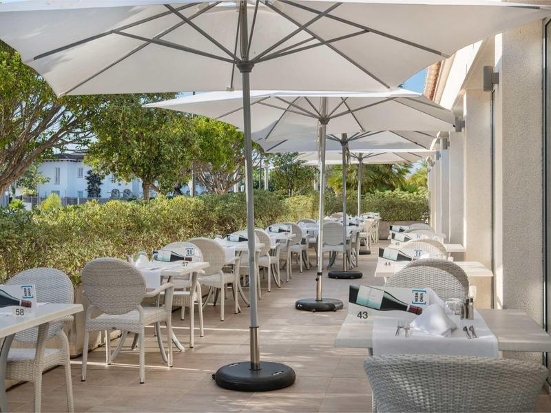 Taras hotelowy z białymi stołami pod parasolami, otoczony zielenią.