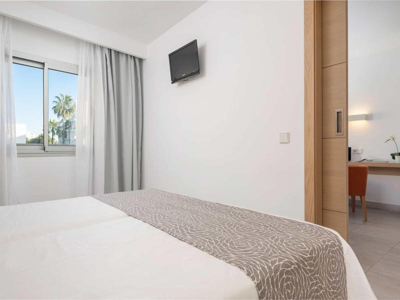 Jasny pokój hotelowy z podwójnym łóżkiem, oknem, szarymi zasłonami i telewizorem na ścianie.