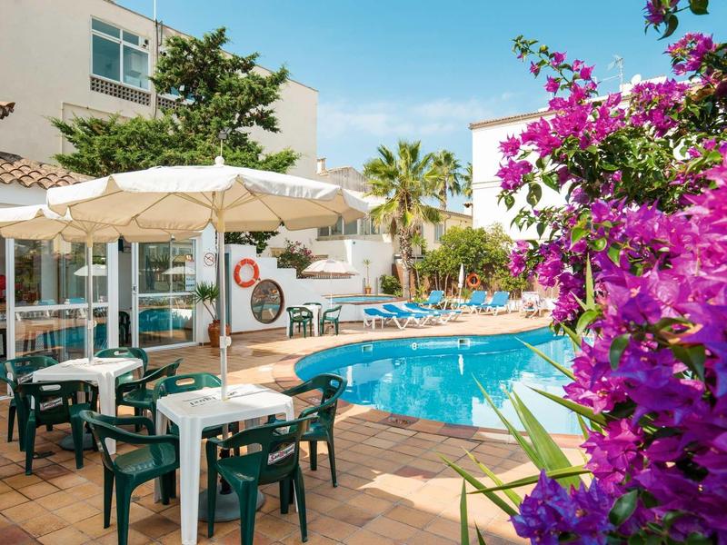 Terrasse d'hôtel avec tables, chaises, parasols et piscine sous un ciel ensoleillé.