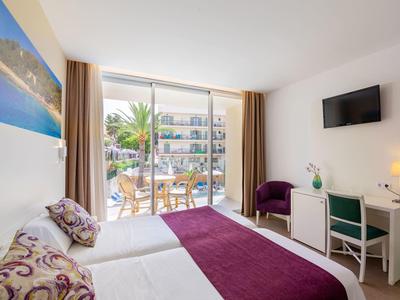 Modern hotel room with double bed, desk, TV, and balcony overlooking garden and buildings.