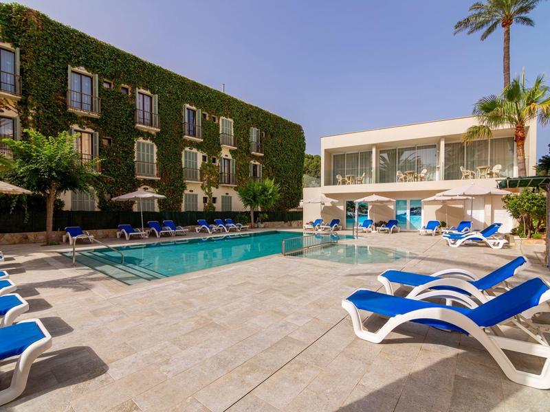 Modern hotel pool with lounge chairs and a green-covered building under a blue sky.