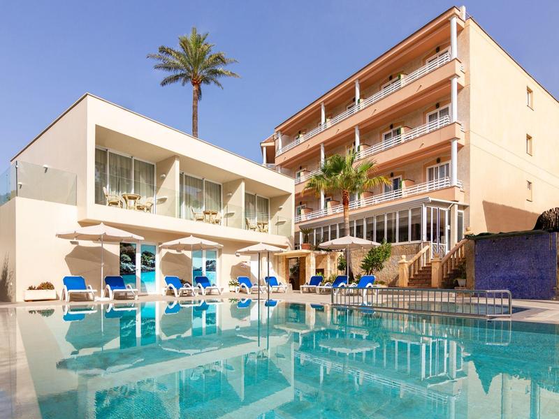 Modern hotel pool with lounge chairs and palm trees beside a light-colored building under a blue sky.
