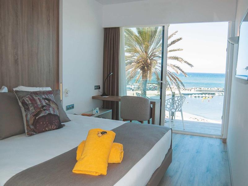 Camera d'hotel moderna con vista mare, letto con asciugamano giallo e balcone con palme.