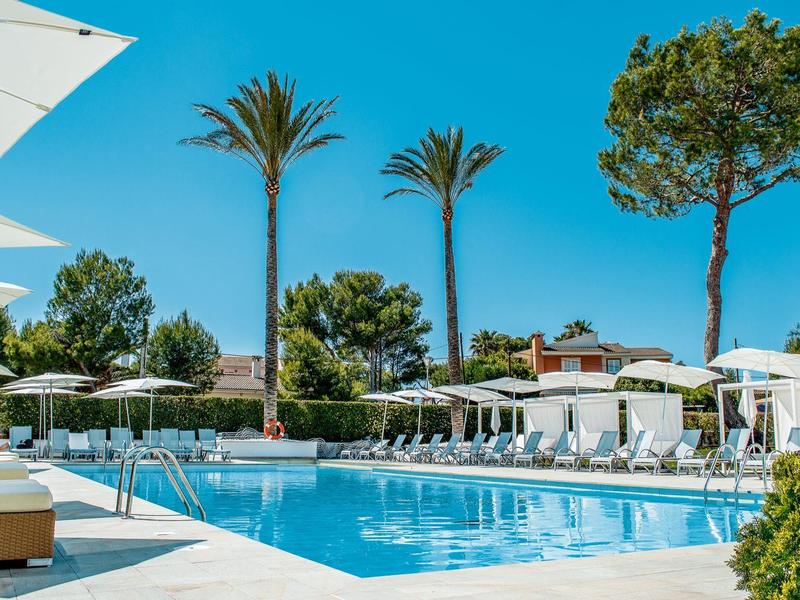 Schwimmbad mit klarem Wasser, Sonnenliegen, weißen Sonnenschirmen und Palmen unter blauem Himmel.