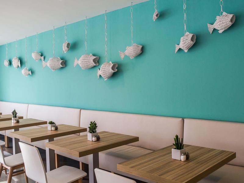 Café mit türkisfarbener Wand, Holztischen, weißen Stühlen und weißen Fisch-Deko an der Wand.