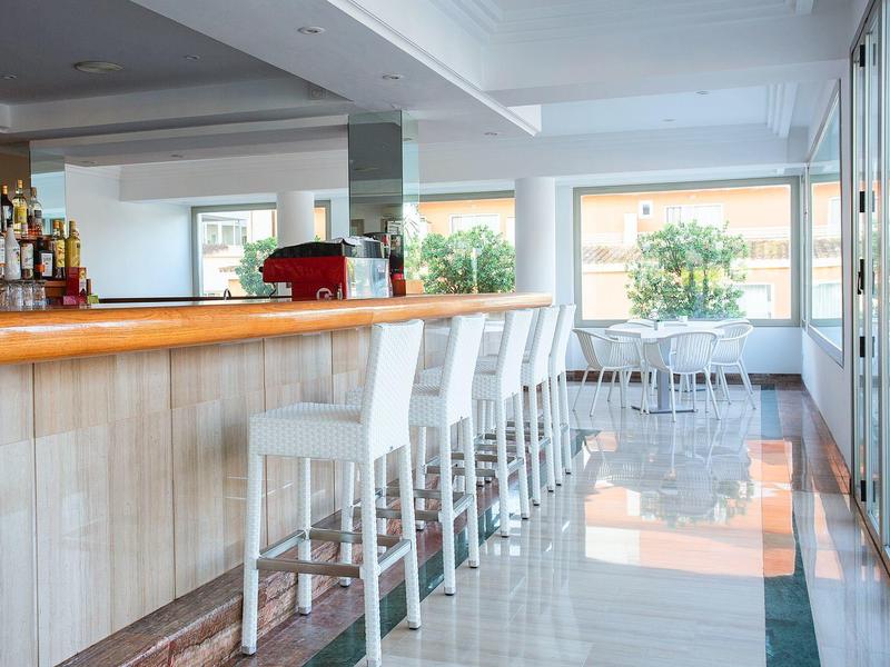 Moderner heller Barbereich mit Holztresen, weißen Barhockern und großen Fenstern mit Pflanzenblick.