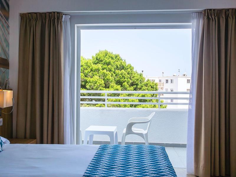 Blick aus hellem Schlafzimmer mit Vorhängen auf Balkon mit weißen Stühlen und grünem Baum im Hintergrund.