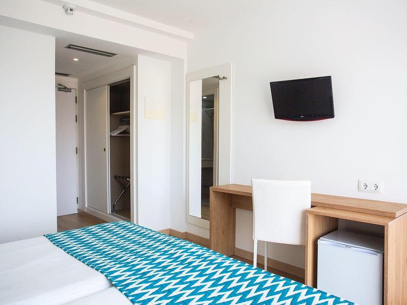 Helles Schlafzimmer mit weißem Bett, blau-weiß gemustertem Bettläufer, Schreibtisch, Stuhl und Wandfernseher.