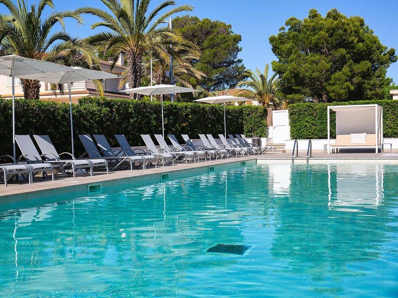 Großer blauer Pool mit hellen Liegestühlen, Sonnenschirmen und Palmen im Hintergrund.