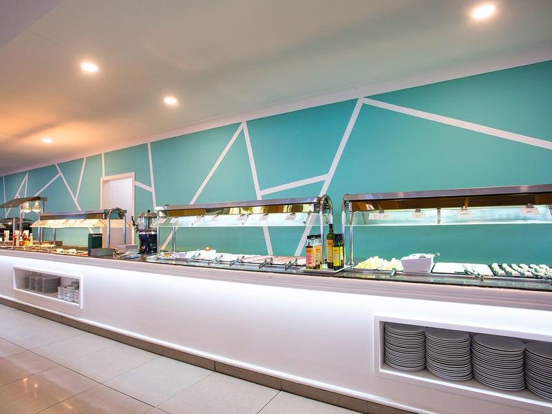 Moderne Buffet-Theke mit türkiser Wand, weißen Linien und beleuchteter Glasvitrine in hellem Raum.