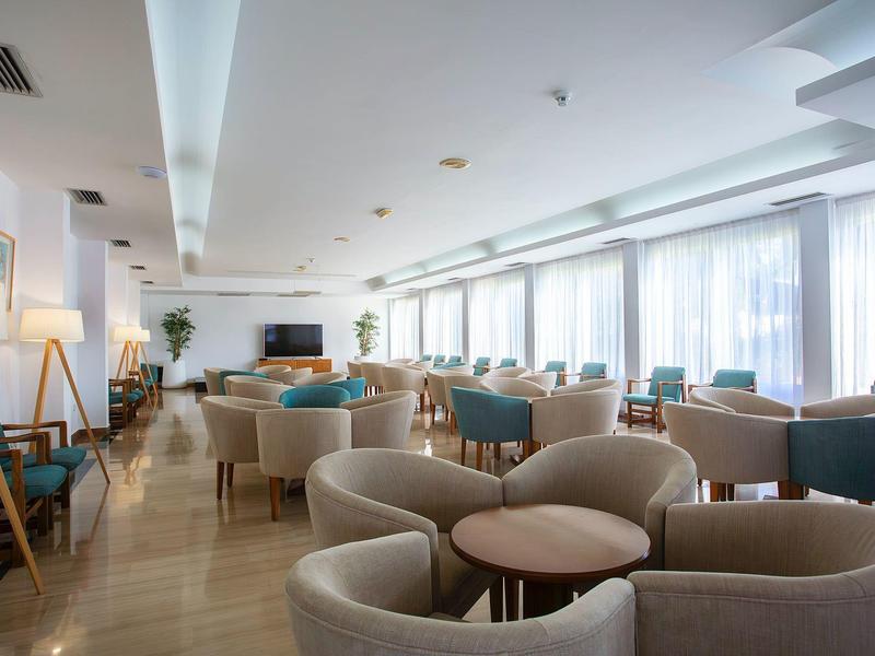 Heller, moderner Lounge-Bereich mit Teppichboden, runden Tischen und beigen Sesseln. Große Fenster.