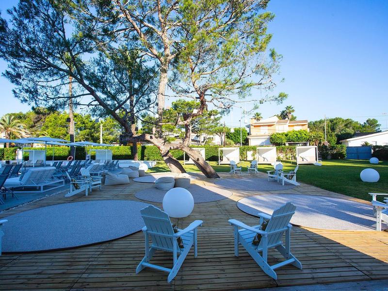 Moderner Außenbereich mit Holzterrasse, weißen Stühlen, grünen Bäumen und klarem blauem Himmel.