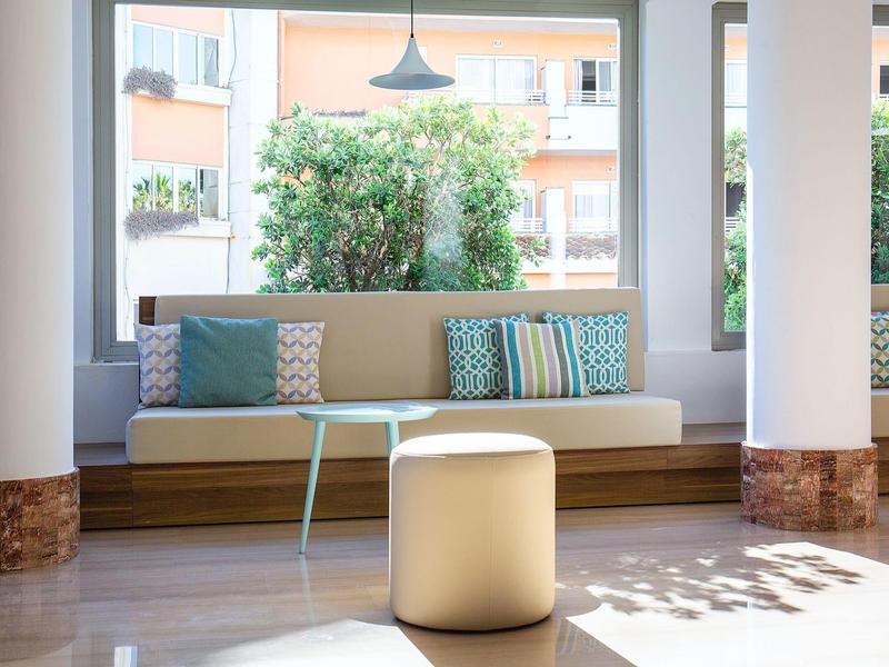 Moderner Wohnbereich mit langem Sofa, gemusterten Kissen und rundem Hocker vor großen Fenstern.