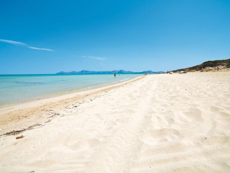 Breiter Sandstrand mit hellem Sand, ruhigem blauem Wasser und klarem Himmel in der Ferne.