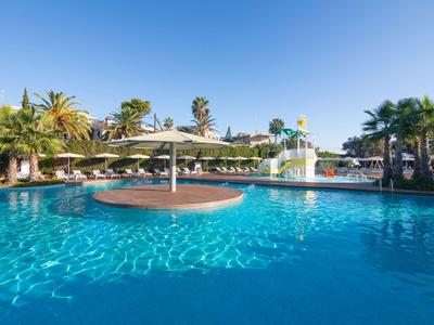 Großer blauer Pool mit Sonnenschirmen, Palmen und Wasserrutsche unter klarem Himmel.