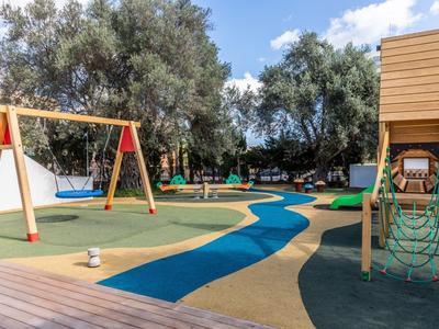 Kolorowy plac zabaw z huśtawkami, drabinką i wybrukowanymi ścieżkami pod drzewami.