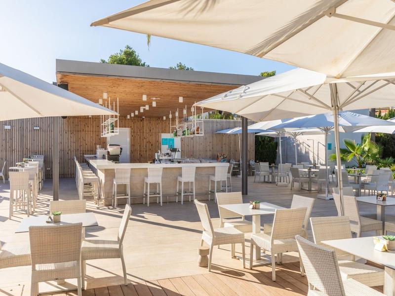 Restauracja na świeżym powietrzu z białymi stołami, krzesłami i dużymi parasolami w ładnej pogodzie.